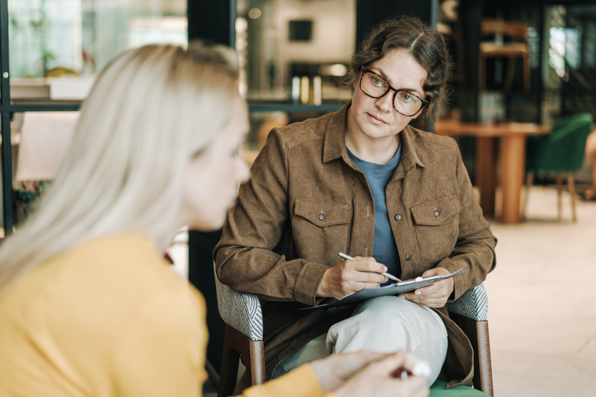 Frau mit Brille und Schreibblock hört einer anderen Frau zu, beide sitzen in einem modernen Raum.