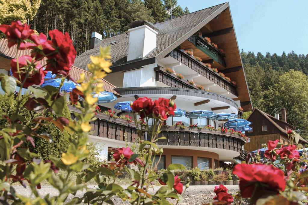 Mehrstöckiges Gebäude mit Balkonen und Sonnenschirmen, umgeben von roten Rosen und Wald im Hintergrund.