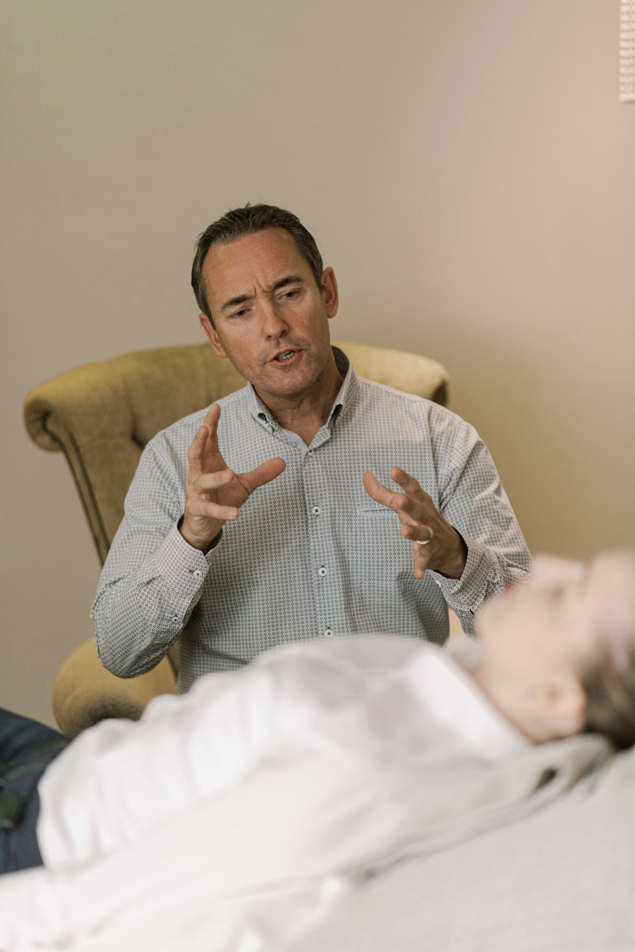 Therapeut im Gespräch mit Patientin auf Liege, ruhige Umgebung.