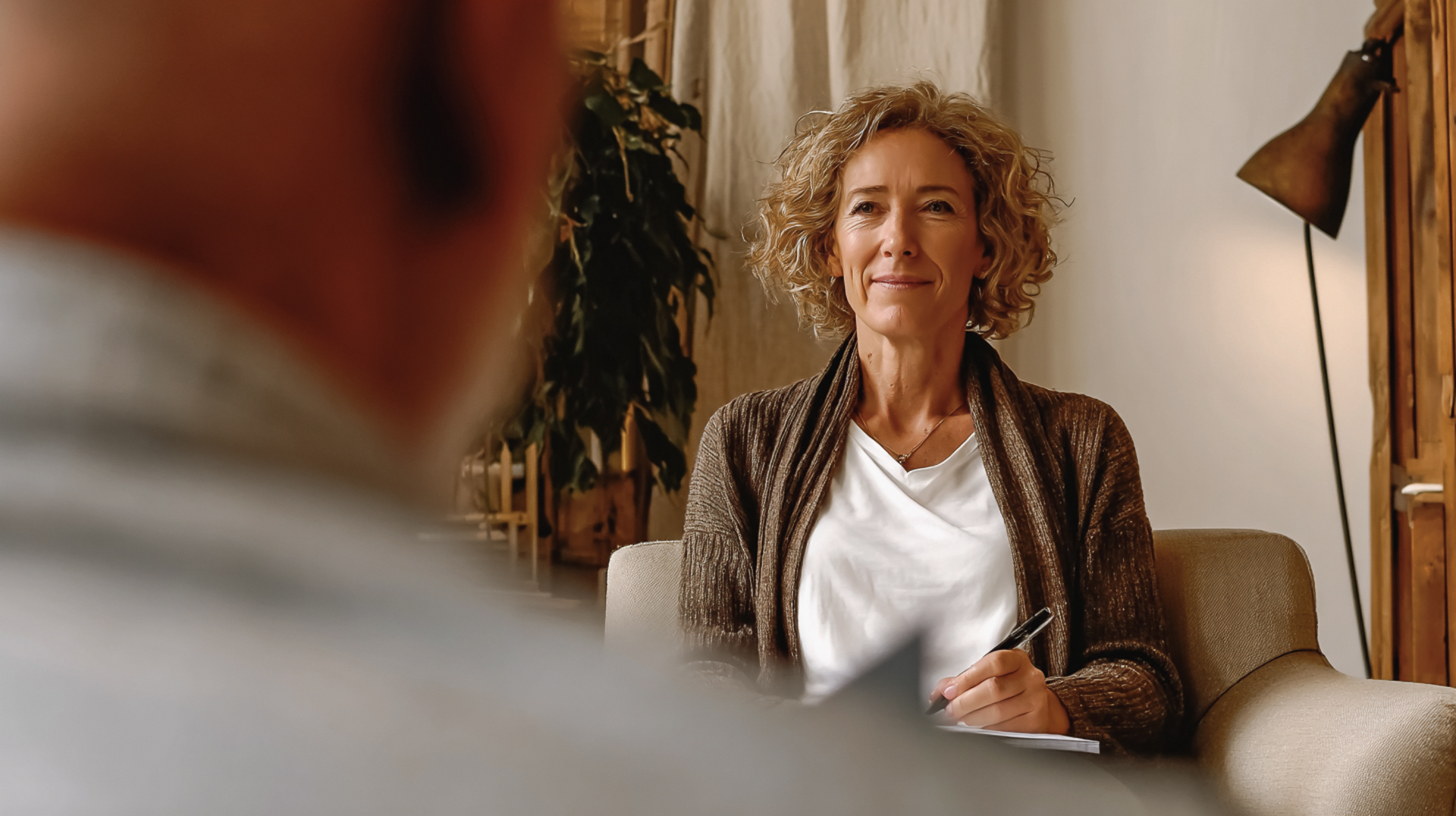 Therapeutin im Gespräch mit Patient, lächelnd, in gemütlichem Raum mit Pflanze und Lampe.