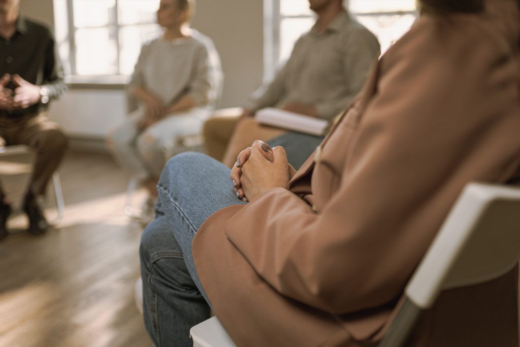 Nahaufnahme einer Gruppentherapiesitzung, Personen auf Stühlen, Fokus auf gefaltete Hände.