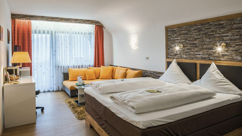 Gemütliches Hotelzimmer mit Doppelbett, orangener Couch und Schreibtisch vor Fenster mit Vorhängen.